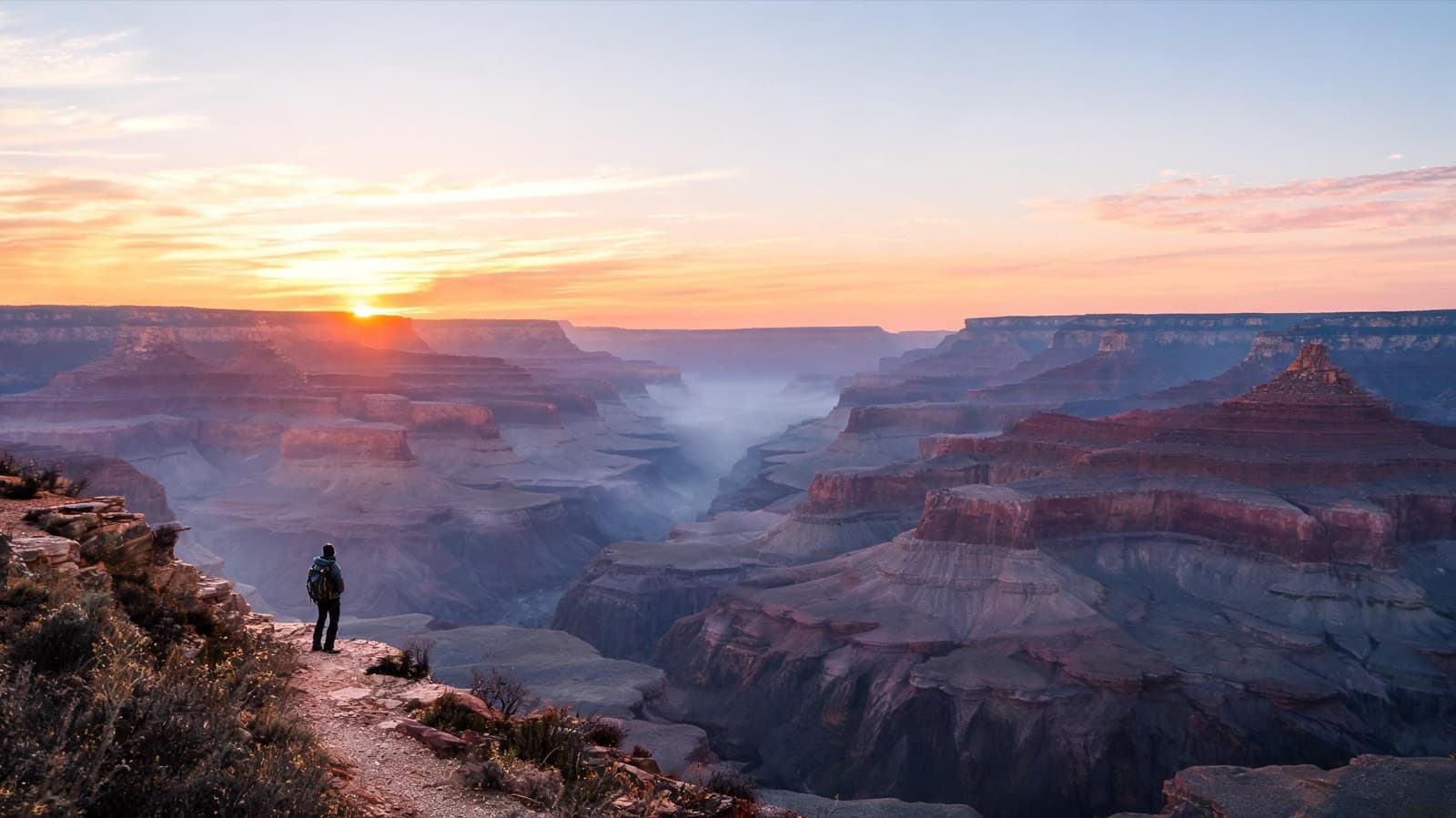 Hiker on a ridge trail overlooking a vast canyon at sunrise, small figure in a wide landscape