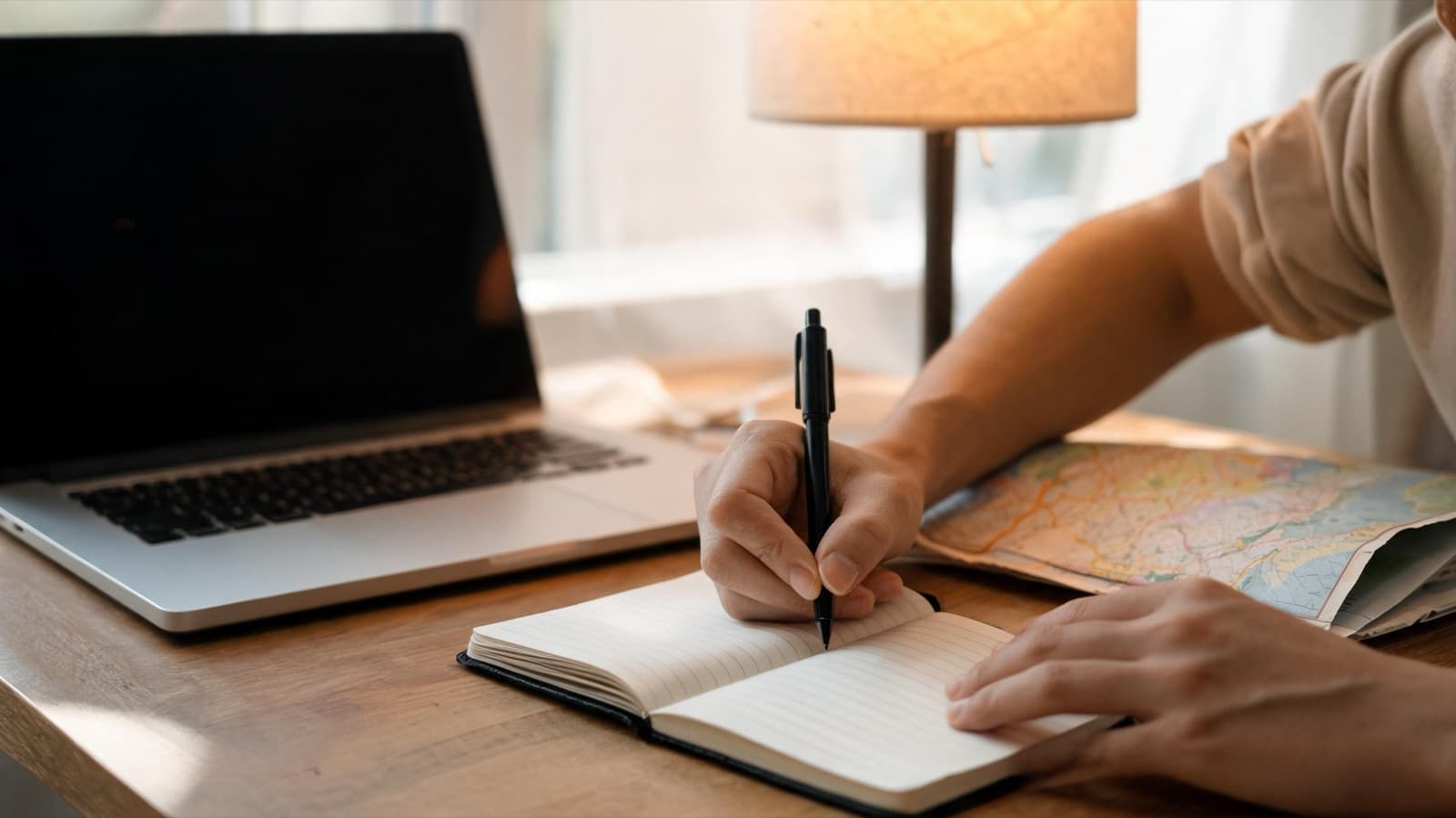 Close-up of hands writing in a travel notebook beside a laptop and a paper map under a warm desk lamp