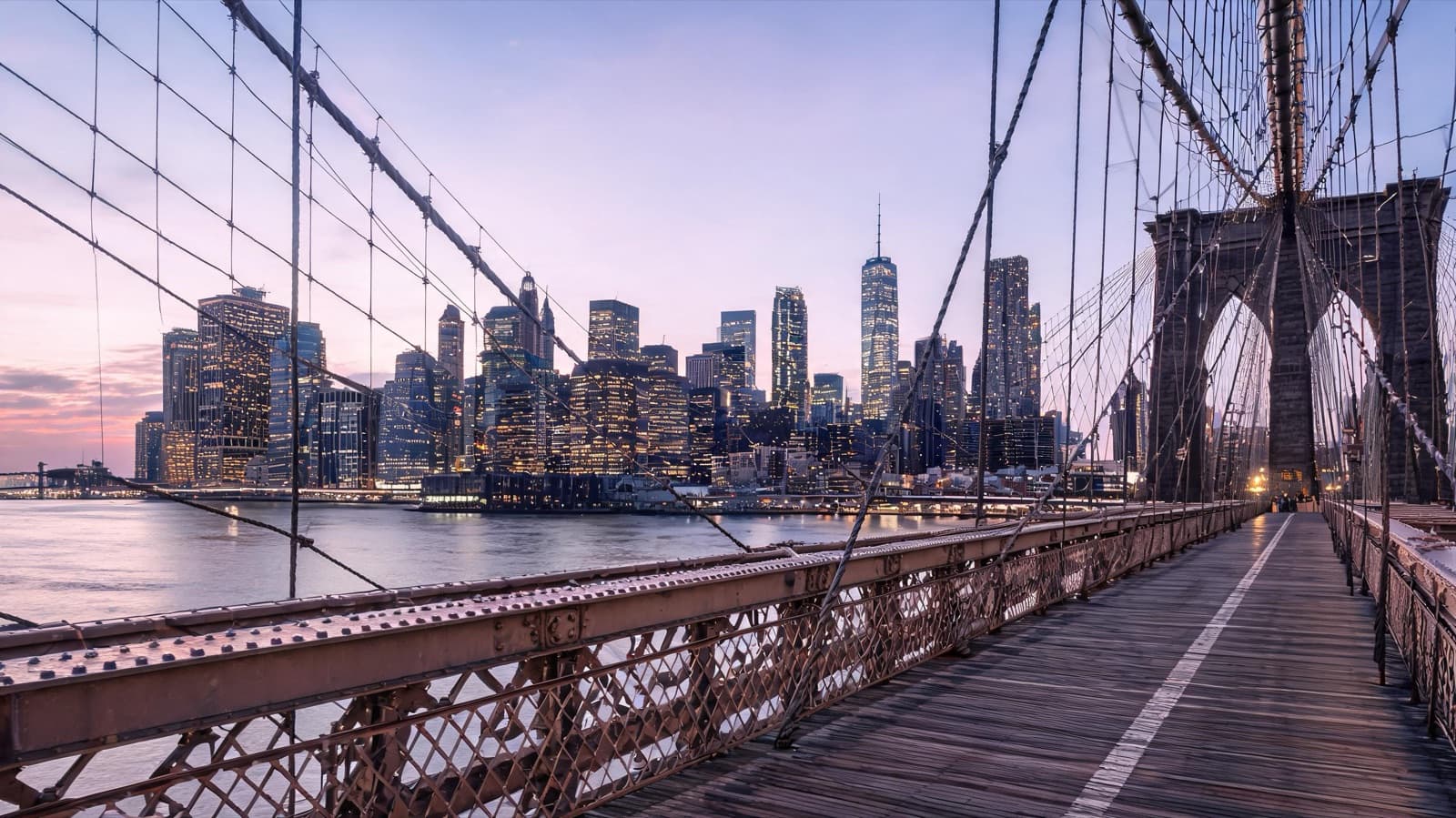 Manhattan skyline at dusk seen from the Brooklyn Bridge walkway, city lights coming on