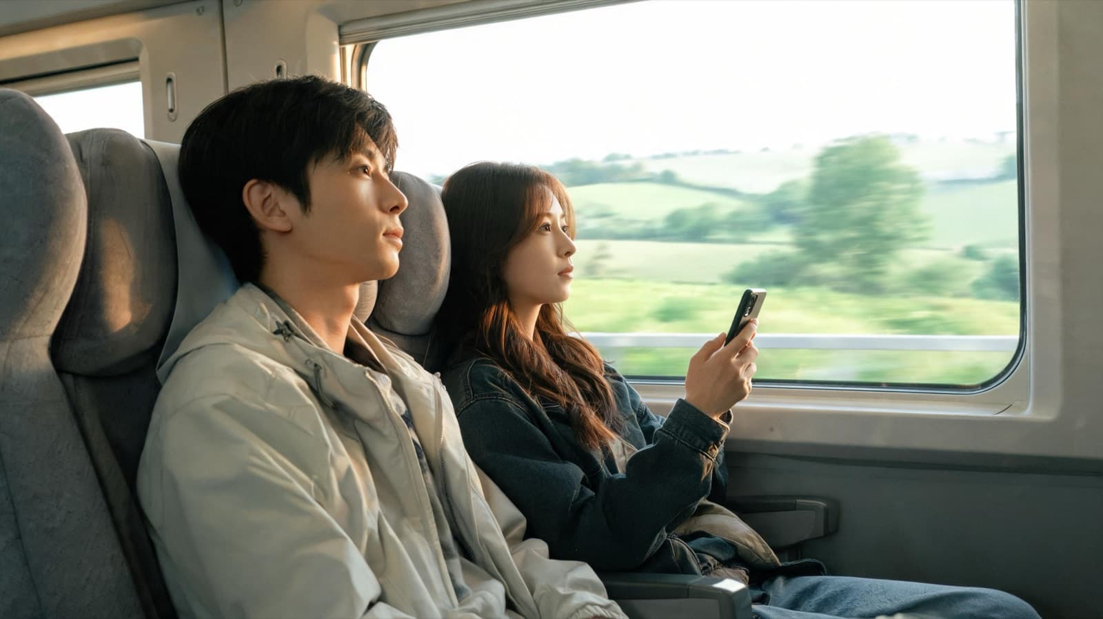 Two travelers by a train window, one holding a phone, landscape blurred outside, screens not visible
